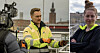Två Mälarenergi-anställda i reflexkläder utomhus med filmkamera och stadsmiljö i bakgrunden.