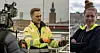 Två Mälarenergi-anställda i reflexkläder utomhus med filmkamera och stadsmiljö i bakgrunden.